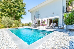 A swimming pool with a white tiled deck, surrounded by a pebble garden, and a white building with a balcony. - Villa Gabrielle - rez-de-jardin, piscine privée (Bassussarry)