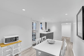 A modern kitchen with white cabinetry, a microwave on a wooden shelf, and a dining table with a bowl. - Modern & Minimalist 2-Bed Suite Near UBC (Vancouver)