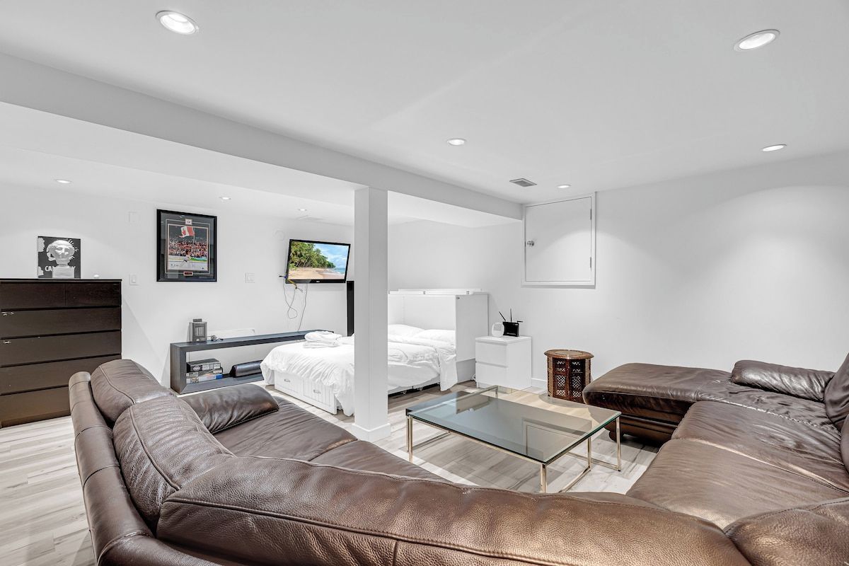 A modern living room with a brown leather sofa, a glass coffee table, and a white bed in the background.