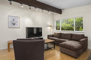 Sala de estar moderna com televisão de tela plana, sofá de couro marrom, mesa de centro de madeira e janela com vista para área verde.