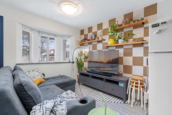Ett vardagsrum med en soffa, en tv, ett soffbord och en vägghylla med växter.