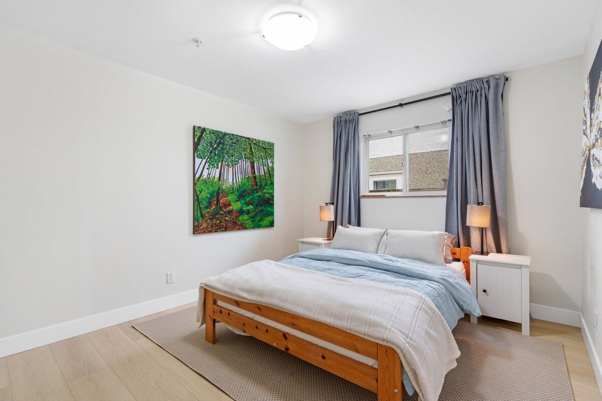 A bedroom with a wooden bed, a nightstand, a lamp, a window with curtains, and a painting on the wall.