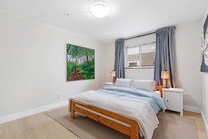 A bedroom with a wooden bed, a nightstand, a lamp, a window with curtains, and a painting on the wall. - Bright & Comfy 1BR Suite | Vancouver’s Prime Area (Vancouver)