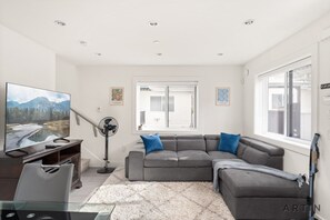 A modern living room with a grey sectional sofa, a flat-screen TV, a glass coffee table, and a large window with blinds. - Sleek Coach House - Designer Kitchen - Sleeps 7 (Vancouver)