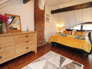 A bedroom with a wooden dresser, a bed with a yellow bedspread, a brick wall, and a window with curtains. - The Thatched Cottage (Preston)