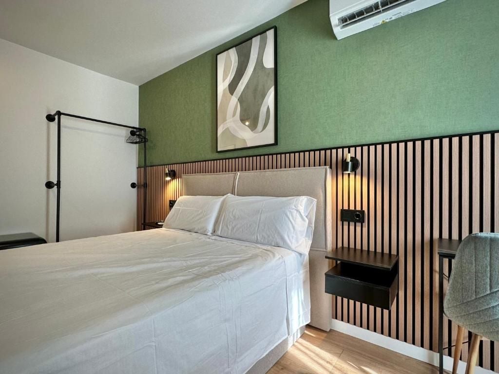 A hotel room with a bed, a wall-mounted air conditioner, a bedside table, and a chair.
