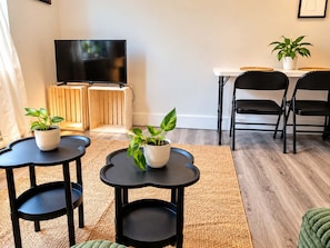 Ein modernes Wohnzimmer mit einem Flachbildfernseher, zwei Blumentöpfen, einem kleinen runden Tisch und einem Essbereich mit Stühlen.