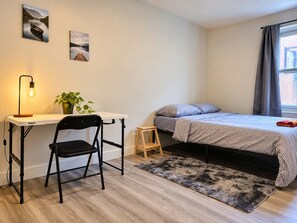 A bedroom with a bed, a desk with a chair, a small stool, a potted plant, and artwork on the wall. - Big 2 rooms appartement 5mn from metro cote ste catherine / pet friendly (Montréal)