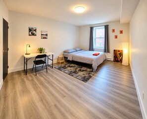Ein Schlafzimmer mit Bett, Schreibtisch mit Stuhl, einer kleinen Hocker, einem Teppich und einem Fenster mit Vorhängen.