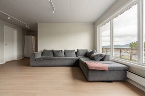 Ein modernes Wohnzimmer mit einer grauen Eckcouch, Holzboden, einem großen Fenster mit Aussicht und einer weißen Decke.