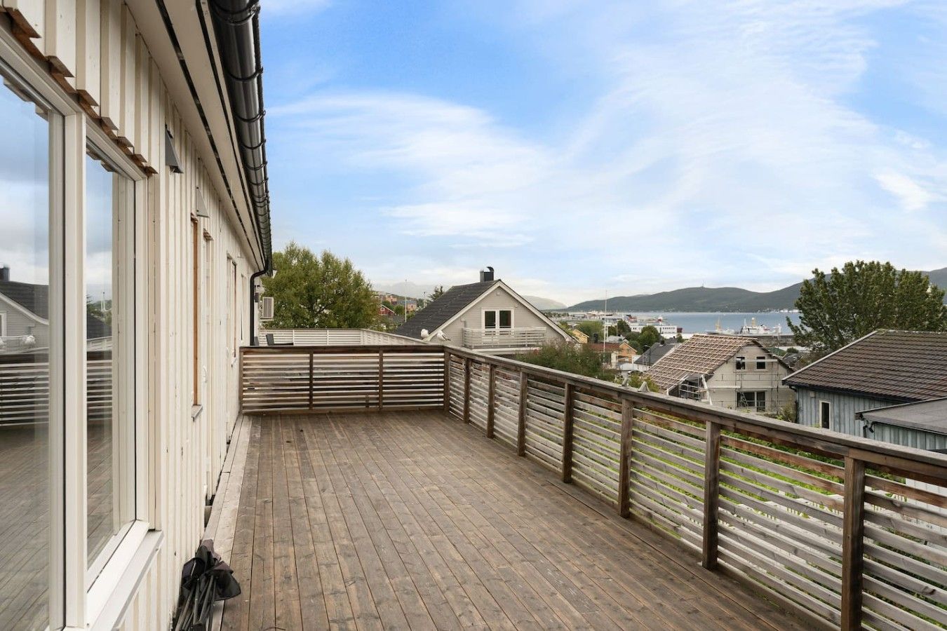 A wooden deck with a view of a lake and hills, surrounded by buildings.