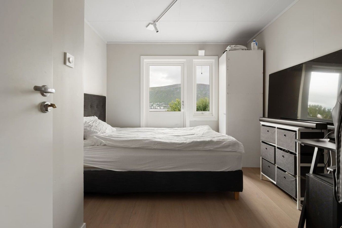 A bedroom with a bed, a dresser, a television, and a window with a view of greenery.