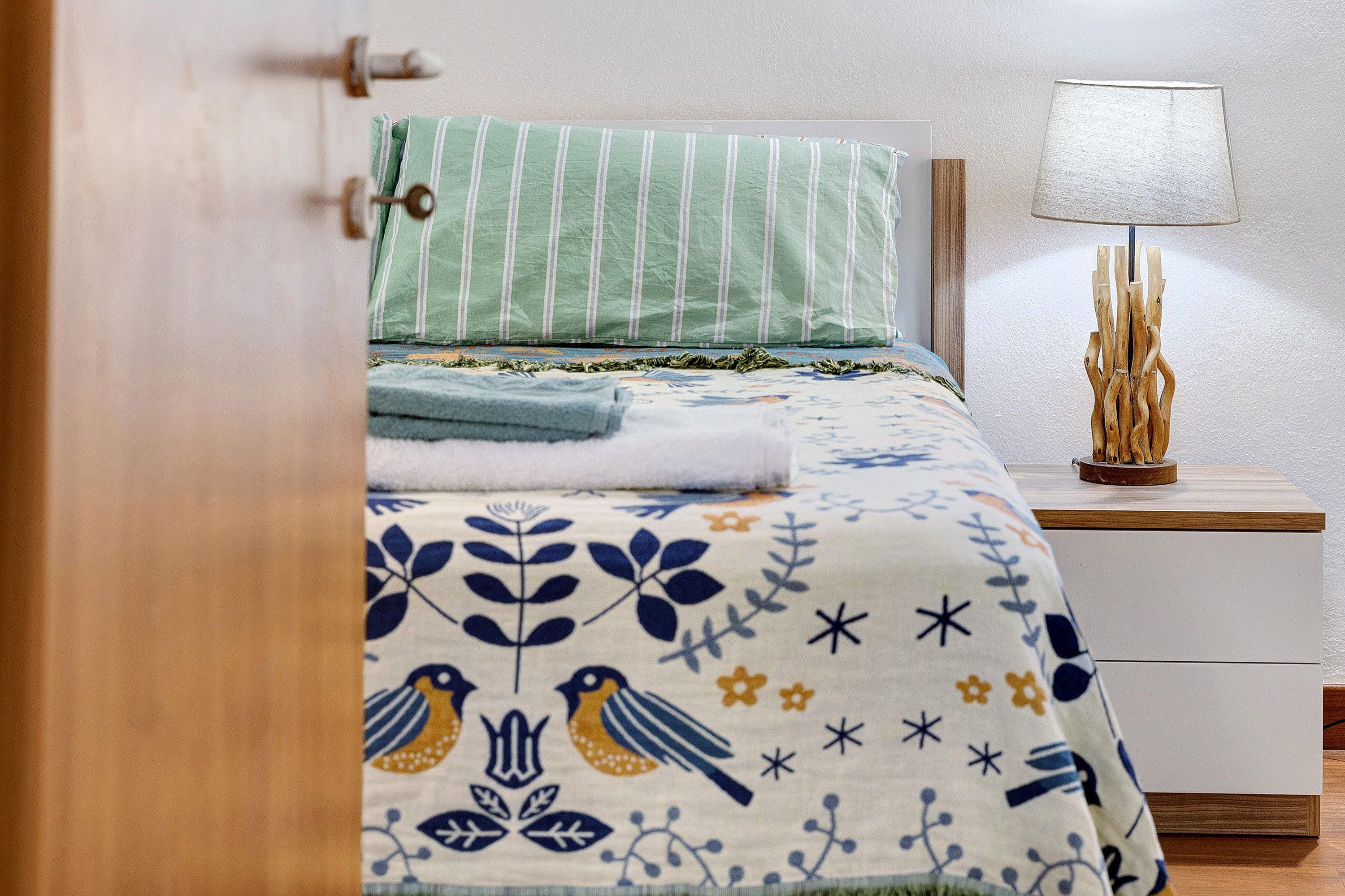 A bed with a striped pillow and a floral-patterned bedspread, a bedside lamp, and a wooden nightstand with a decorative item.