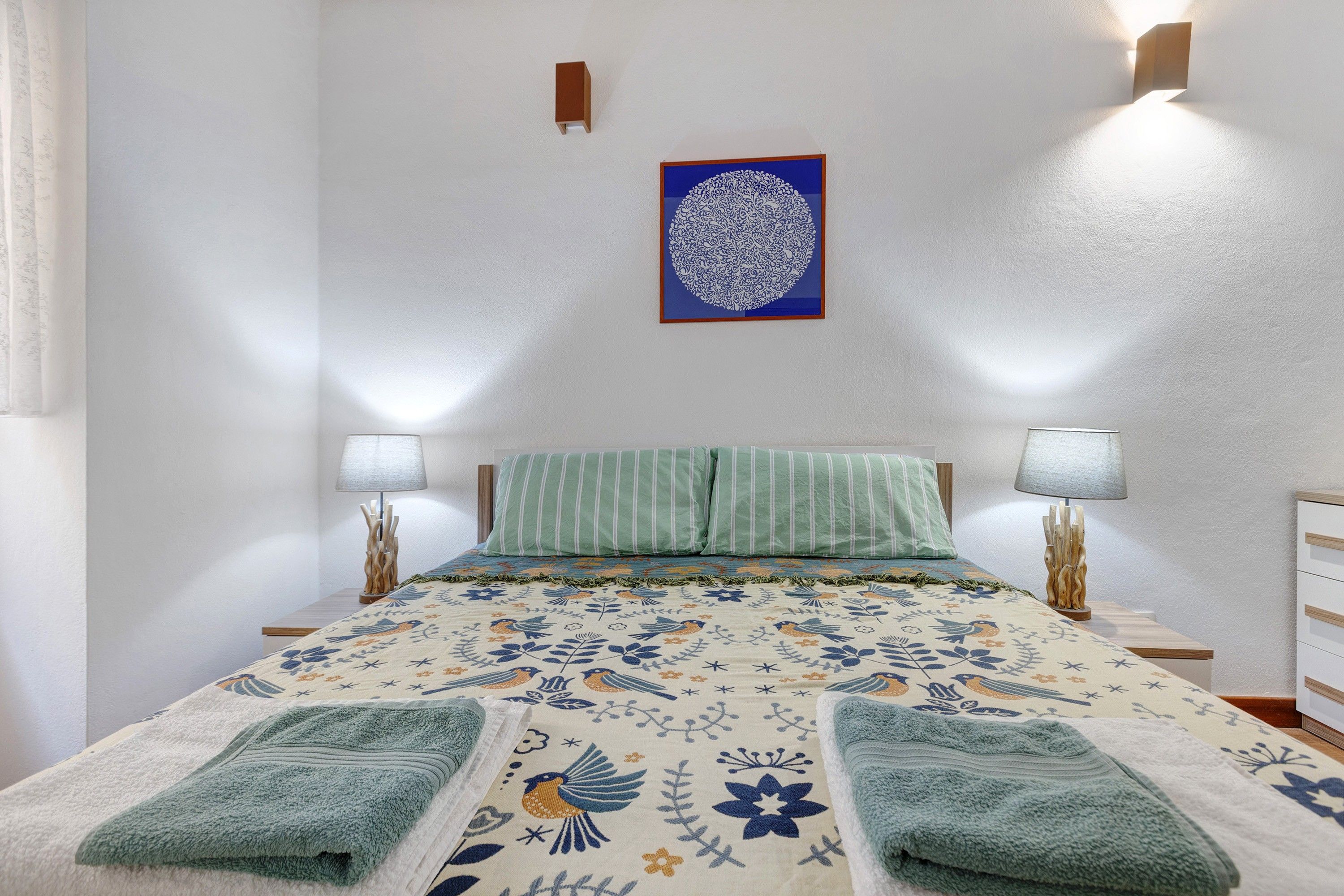 A bedroom with a bed, two bedside lamps, a wall-mounted light fixture, and a framed artwork.