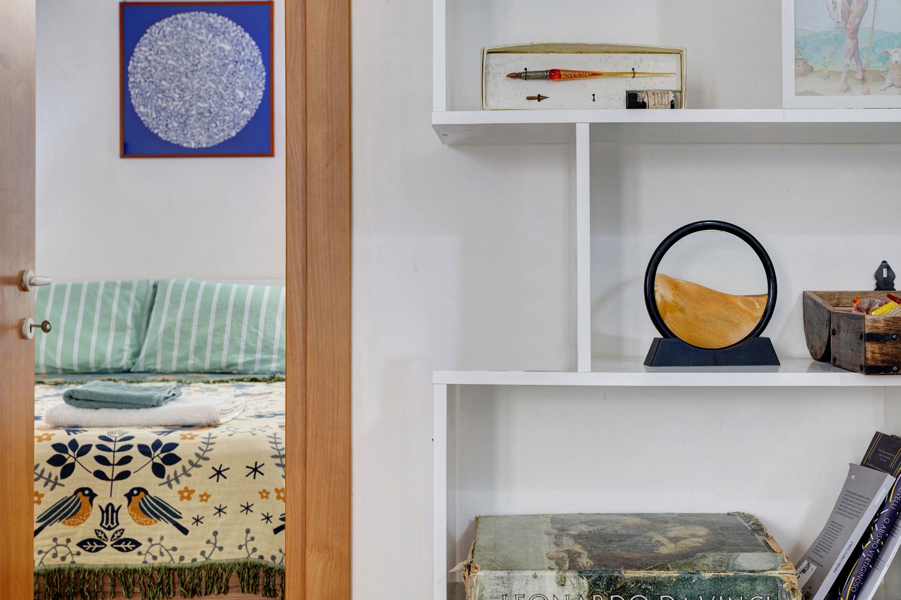 A room with a bed, a shelf with books, a framed picture, and a small table with a lamp.