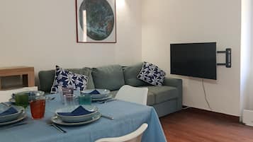 Een eetgedeelte met een tafel gedekt voor twee personen, een bank met gestreepte kussens, een wandgemonteerde flatscreen-tv en een kunstwerk in een lijst aan de muur.