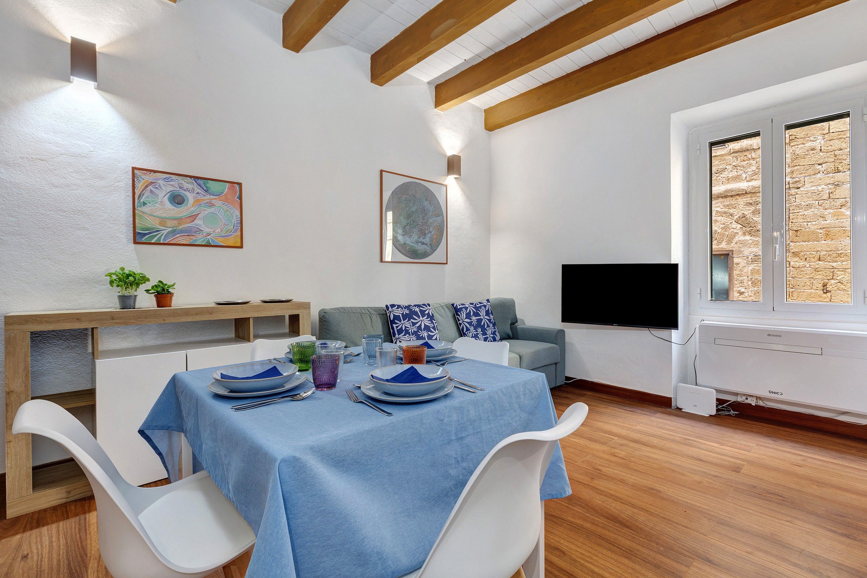 A dining area with a table set for four, a sofa, a TV, and a window with a view of stone walls.