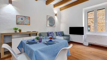 A dining area with a table set for four, a sofa, a TV, and a window with a view of stone walls.