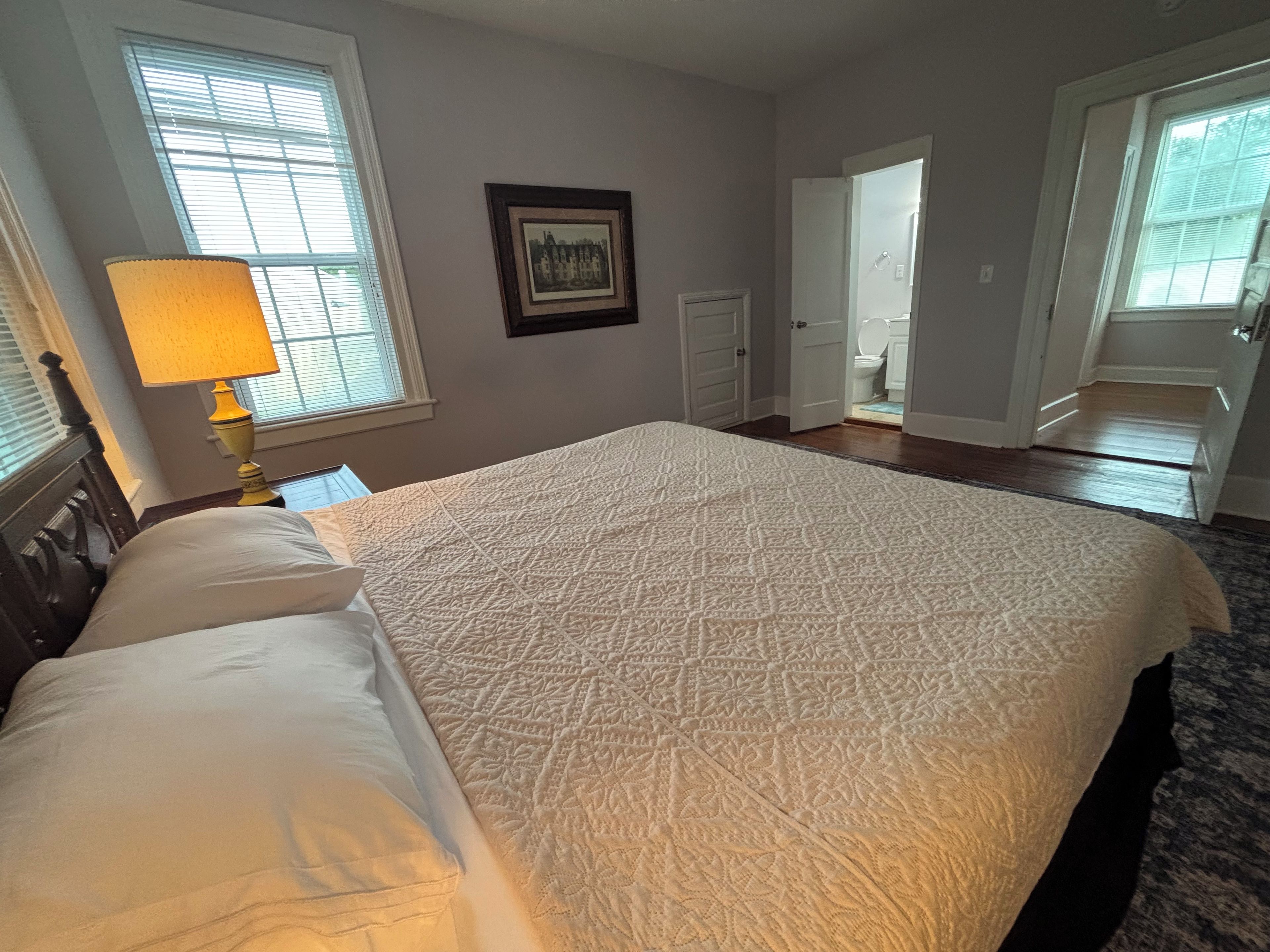 A bedroom with a bed, a bedside lamp, a framed picture on the wall, and a door leading to a bathroom.