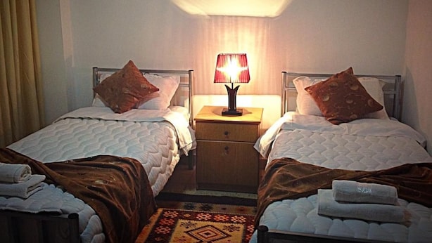 Quarto de hotel com duas camas, uma mesa de cabeceira com abajur e um tapete estampado.