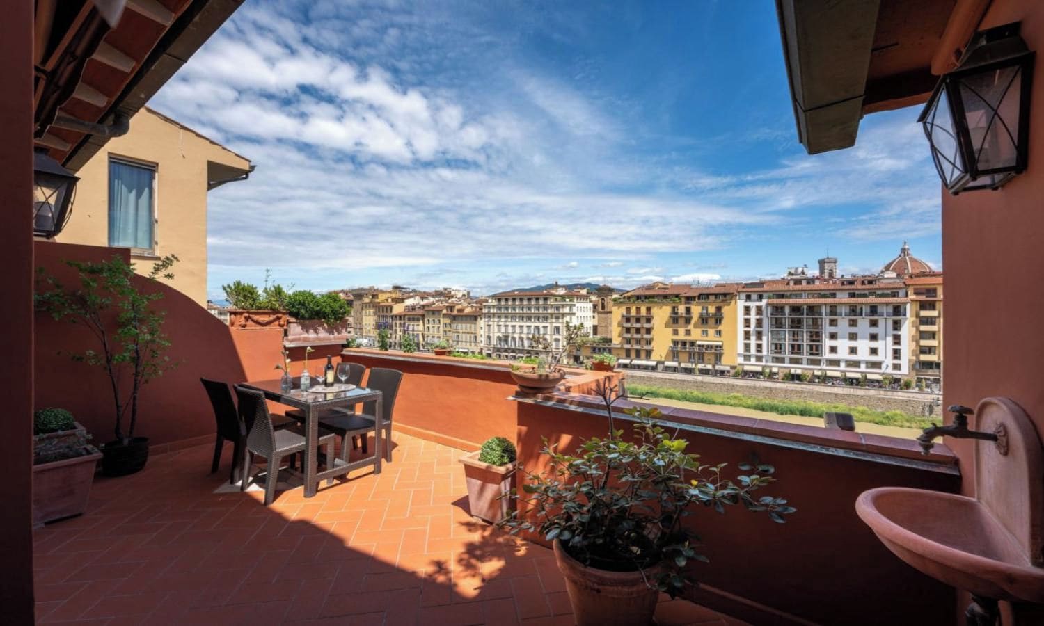 A terrace with a table and chairs, potted plants, and a view of city buildings.