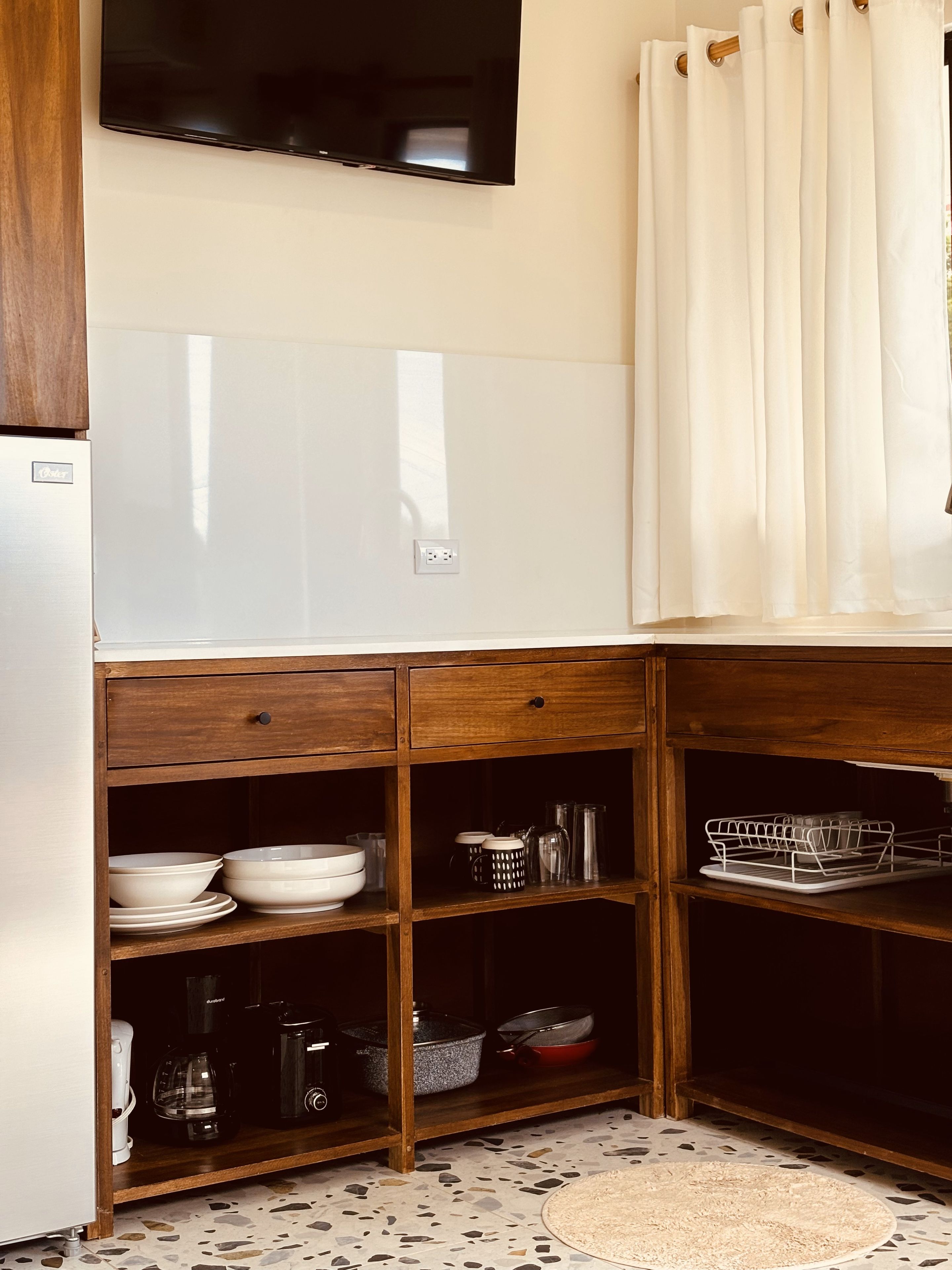 Una cocina con lavavajillas integrado, un televisor de pantalla plana montado sobre la encimera y un armario de madera con estantes abiertos que contienen utensilios de cocina.