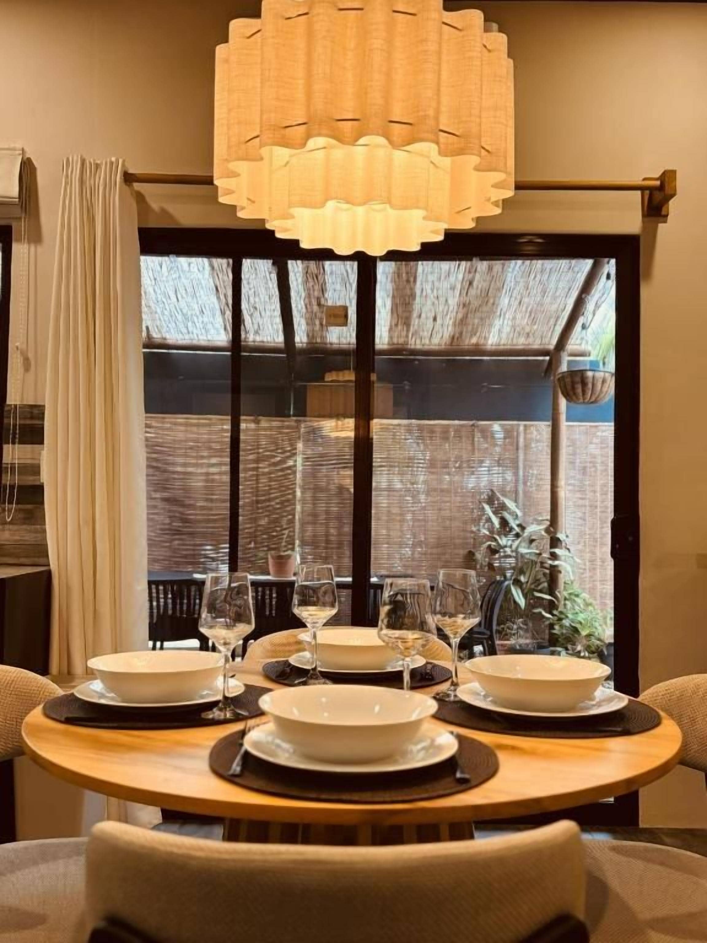 A dining table set for four with white plates and glasses, a hanging light fixture above, and a view through a window with a wooden structure outside.