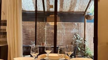 A dining table set for four with white plates and glasses, a hanging light fixture above, and a view through a window with a wooden structure outside.