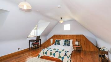 Ein Schlafzimmer mit geneigter Decke, einem Bett mit gemusterten Textilien, einem Schreibtisch mit Stuhl und einem Fenster.