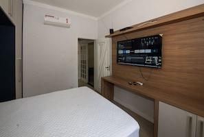 A bedroom with a bed, a TV mounted on a wooden cabinet, and a white air conditioner on the wall. - SHB - 3 min praia - Central - Biquinha (São Vicente)