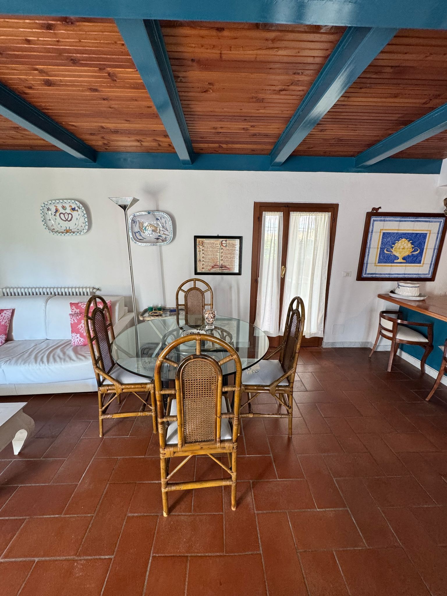 A dining area with a glass table surrounded by wicker chairs, a white sofa with pink pillows, and a blue-framed picture on the wall.