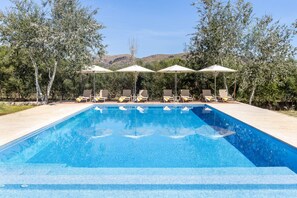 Ein rechteckiger Swimmingpool mit klarem, blauem Wasser, umgeben von Liegestühlen und Sonnenschirmen, vor einer Kulisse aus Bäumen und Hügeln.