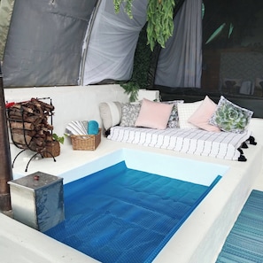 Un área de piscina con una piscina azul, un sofá blanco con cojines, una cesta de mimbre y un recipiente metálico.