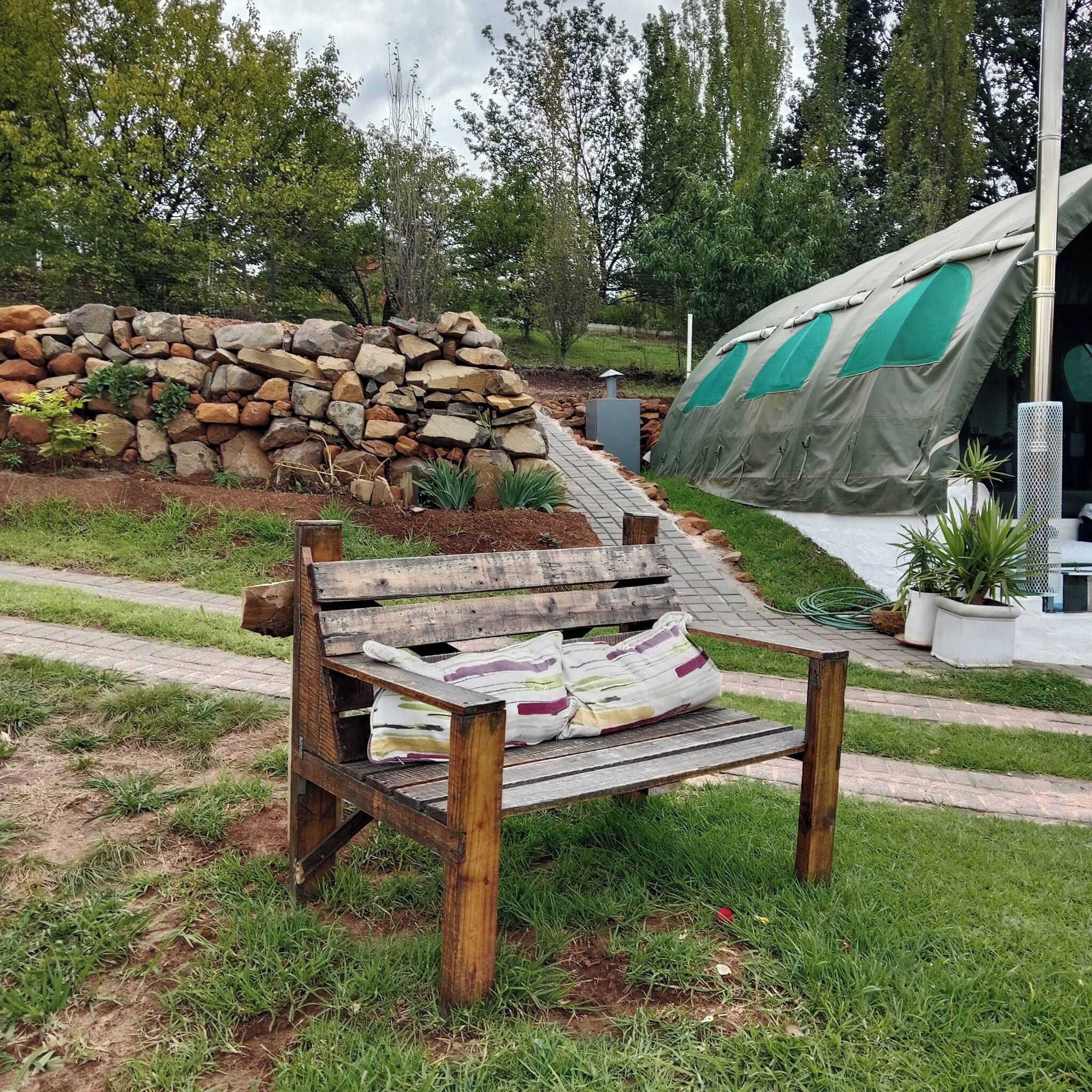 A rustic wooden bench with a cushion, a stone wall, and a green tent in a garden setting.