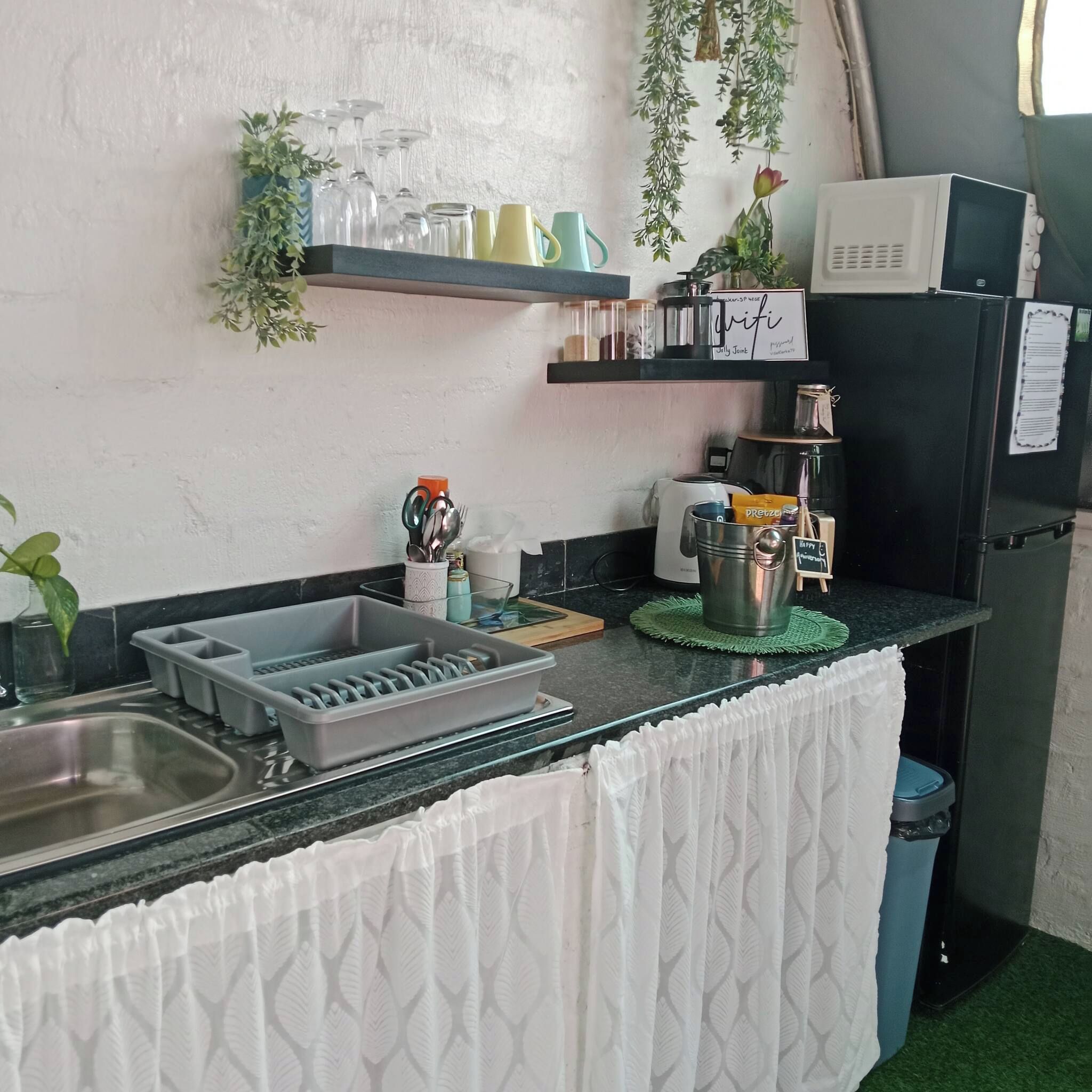 Cocina compacta con lavamanos, microondas y refrigerador. Hay repisas sobre la encimera con varios utensilios de cocina.