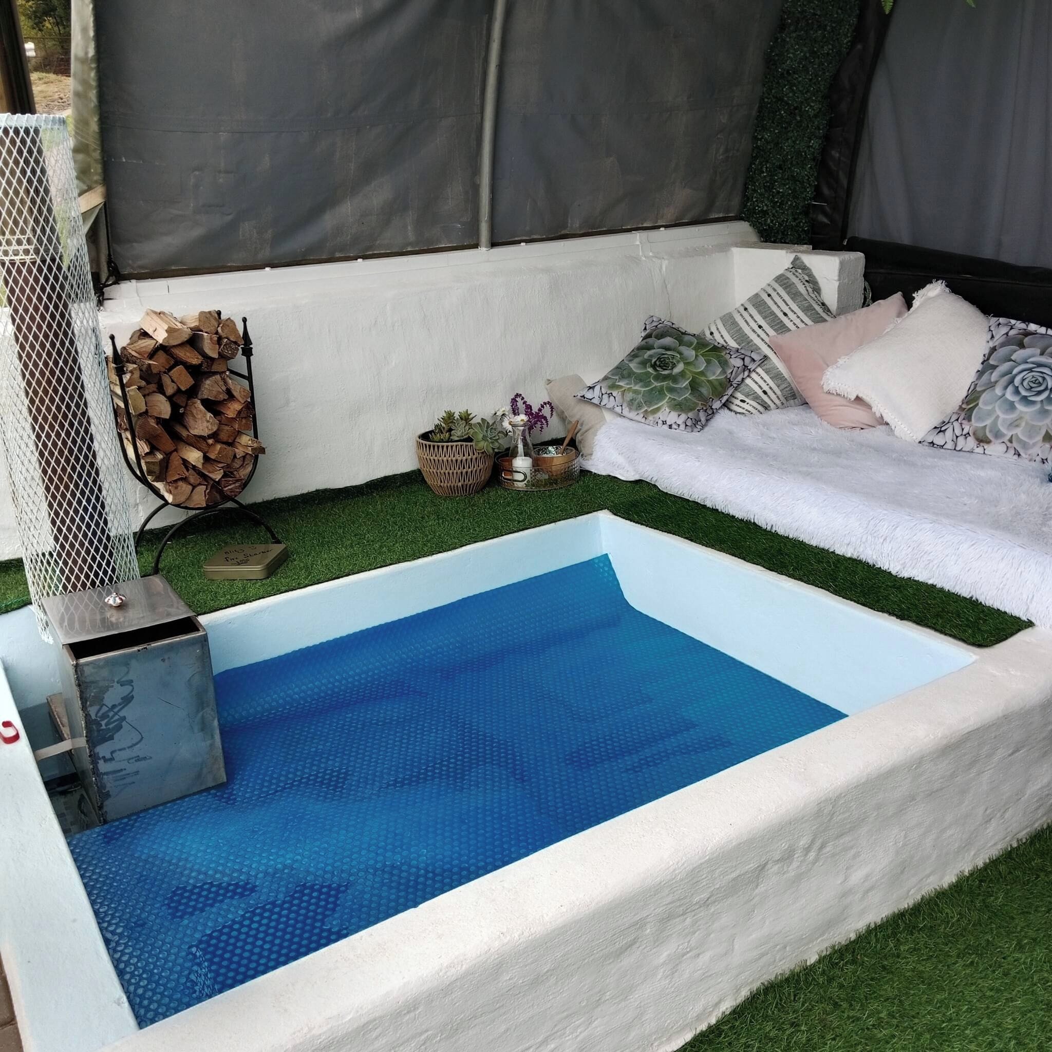 Una pequeña piscina con piso azul texturizado, rodeada por un borde blanco, con una zona verde y un área de descanso con cojines y una planta.