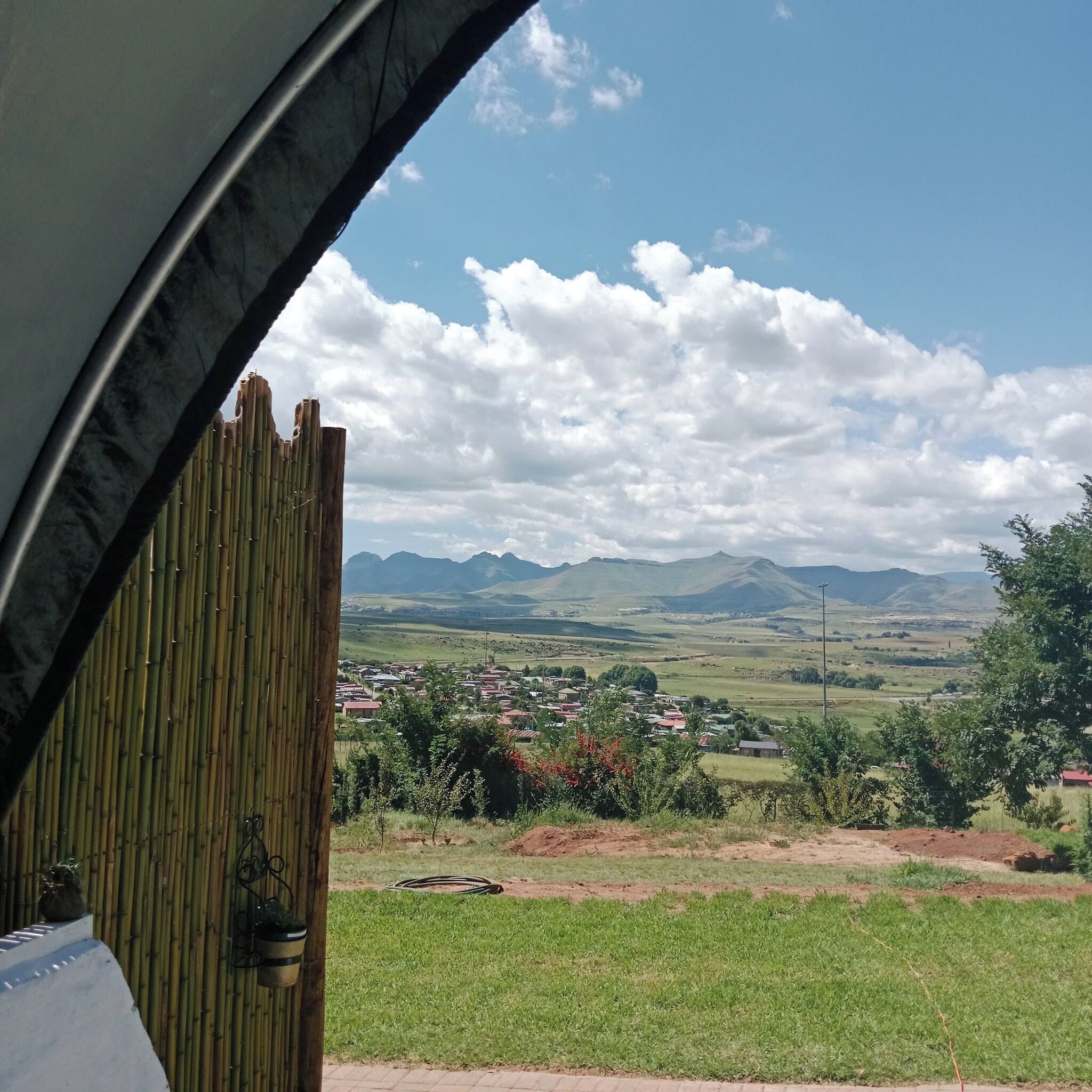 Vista pela janela com persianas de bambu, paisagem rural com montanhas e um pequeno povoado ao longe.