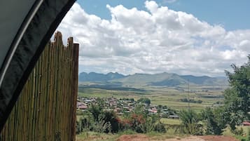 Vista pela janela com persianas de bambu, paisagem rural com montanhas e um pequeno povoado ao longe.