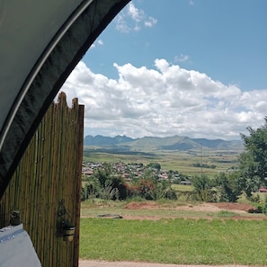 Vista a través de una ventana con persianas de bambú, paisaje rural con montañas y un pequeño asentamiento en la distancia.