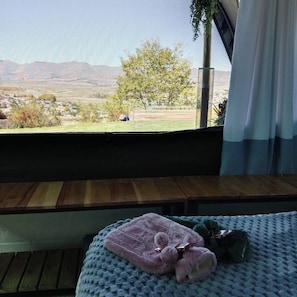 Una habitación con vistas al paisaje, una cama con una manta y un osito de peluche, y una ventana con cortinas.