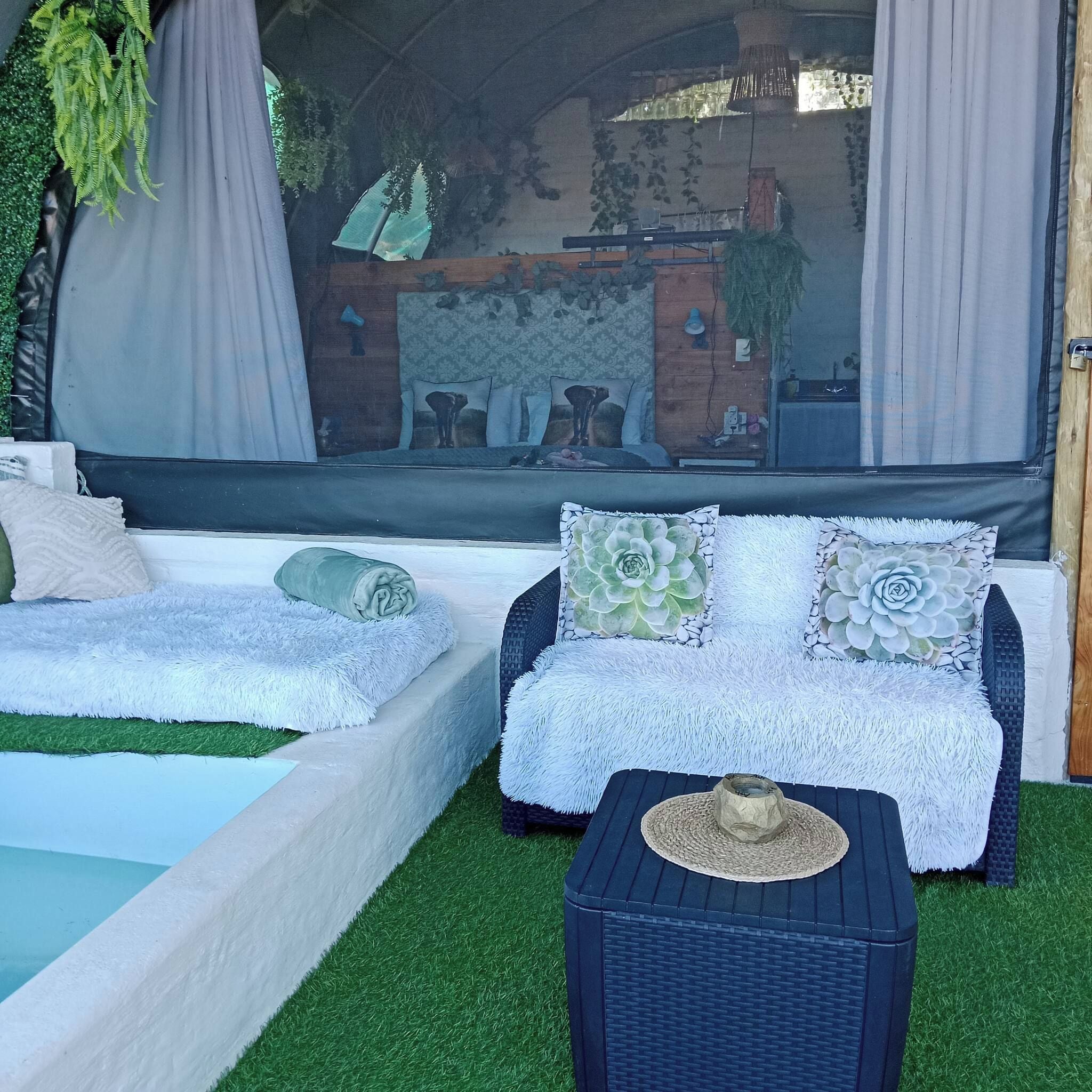 A cozy outdoor seating area with a sofa and a small table, surrounded by artificial grass and a view of a brick wall and greenery.