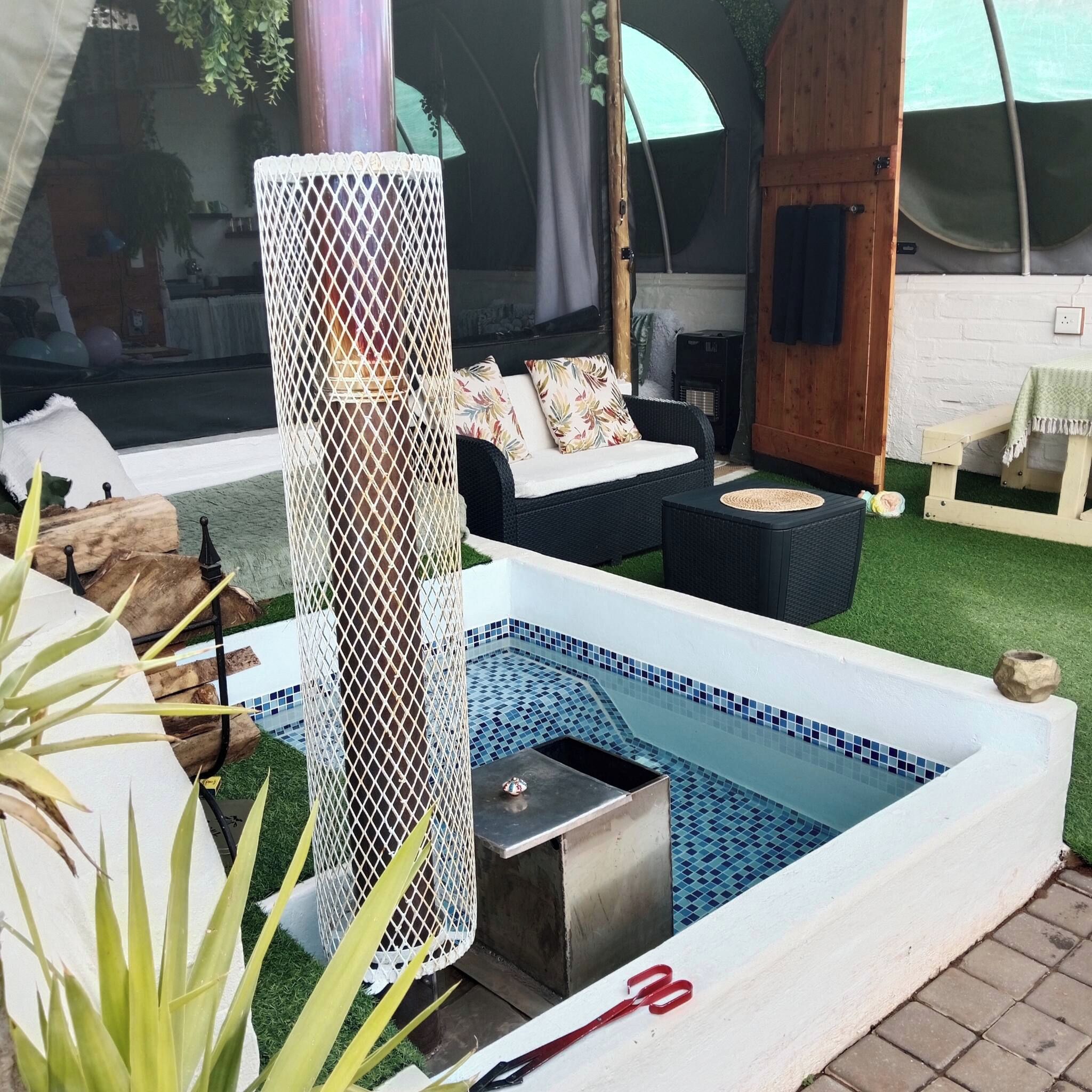 Área de piscina com borda de azulejos brancos, uma banheira de hidromassagem de metal e um sofá de vime com almofadas.