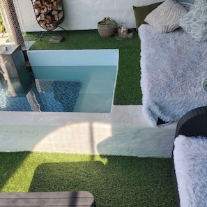 Una piscina con césped artificial, un sofá blanco y mullido, y una cesta de mimbre con una botella.