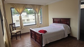 A bedroom with a wooden bed, a chair, a window with curtains, and a view of buildings outside.