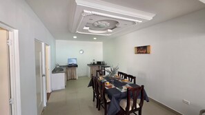 A dining area with a table set for four, a kitchen in the background, and a ceiling with recessed lighting.