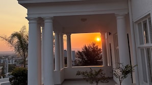 En bygning med en overdækket veranda understøttet af søjler, udsigt til solnedgang og et lille træ i forgrunden.