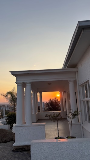 Un bâtiment doté d’un porche couvert soutenu par des colonnes, offrant une vue sur le coucher de soleil, et avec un petit arbre au premier plan.