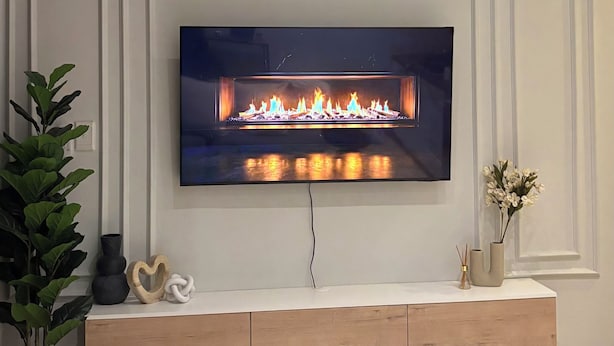 En moderne stue med et fladskærms-tv monteret på væggen, et træunderholdningsmøbel nedenunder og en potteplante til venstre.