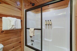 Puisilla paneeleilla verhoiltu kylpyhuone, jossa on lasiseinällä varustettu suihku, pyyhekoukku ja kolme pulloa shampoota.