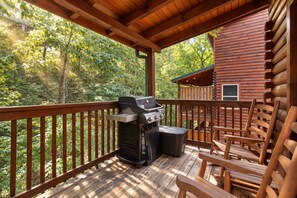 A wooden deck with a grill and chairs, overlooking a forested area. - Creekside Lodge | Hot Tub, Grill, Fireplace, GAMES (Pigeon Forge)